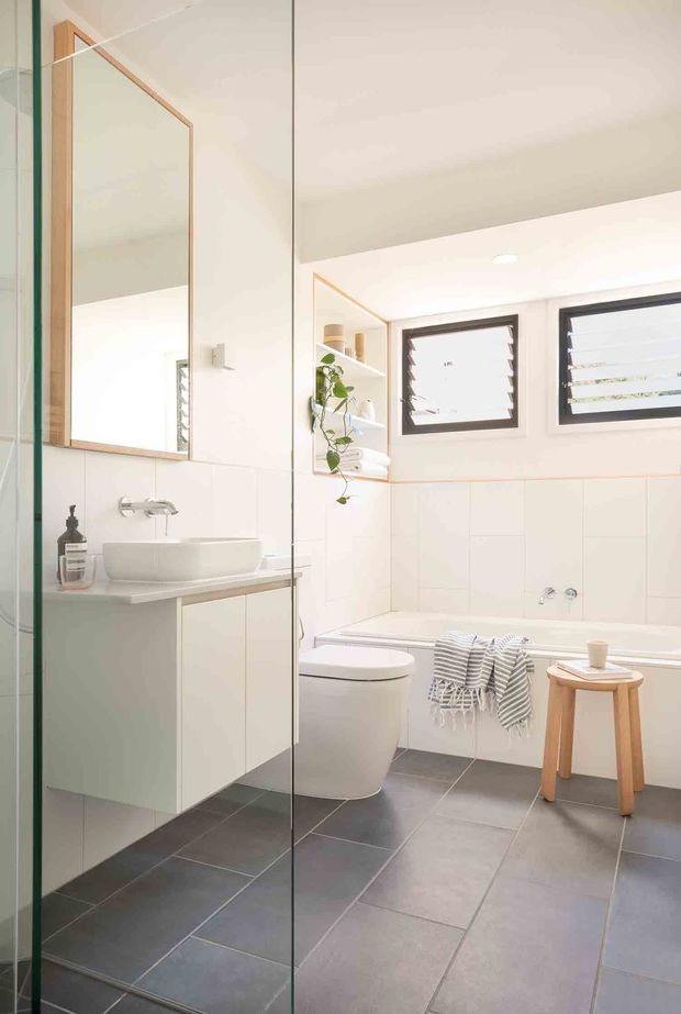 Bright contemporary bathroom featuring a frameless glass shower enclosure, wall-mounted white vanity with integrated basin, large timber-framed mirror, freestanding toilet, bathtub, recessed shelving with greenery, and dark grey floor tiles. High horizontal windows with dark frames and louvred glazing provide ventilation and natural light. The palette is predominantly white with grey flooring and light timber accents.