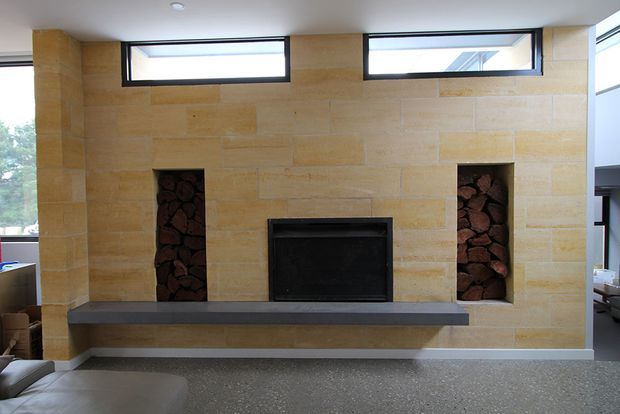 A contemporary living room fireplace feature wall clad in warm beige natural stone, with a black firebox, long dark projecting hearth, and two vertical niches filled with stacked firewood or dark stones. Narrow clerestory windows bring in natural light, while adjacent seating and glazing are partially visible at the sides.