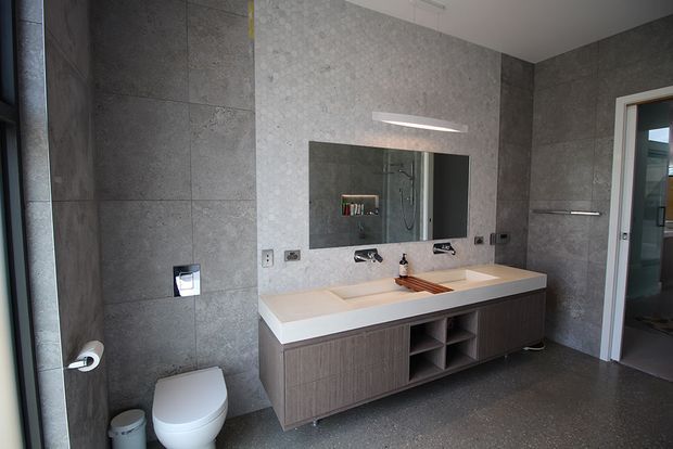 Contemporary bathroom with large-format dark grey wall and floor tiles, a long double vanity with a light stone benchtop and dark wood-grain cabinetry, a wide wall mirror, wall-mounted fittings, and a white toilet. A glass shower enclosure is visible through the mirror and doorway.