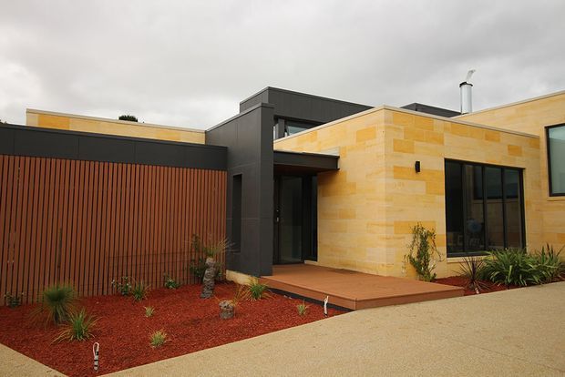 Single-storey contemporary residence viewed from the front, with warm golden stone masonry, dark charcoal cladding and trim, a tall timber slat garage or privacy screen, and a recessed entry. The landscaped frontage uses red gravel, sparse drought-tolerant planting, and a pale paved driveway under an overcast sky.