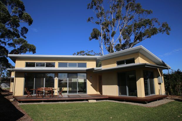 Exterior view of a low-rise contemporary house with warm beige rendered walls, broad horizontal glazing, projecting roof forms, and a timber deck opening onto a lawn. Mature eucalyptus trees and a clear blue sky surround the property.
