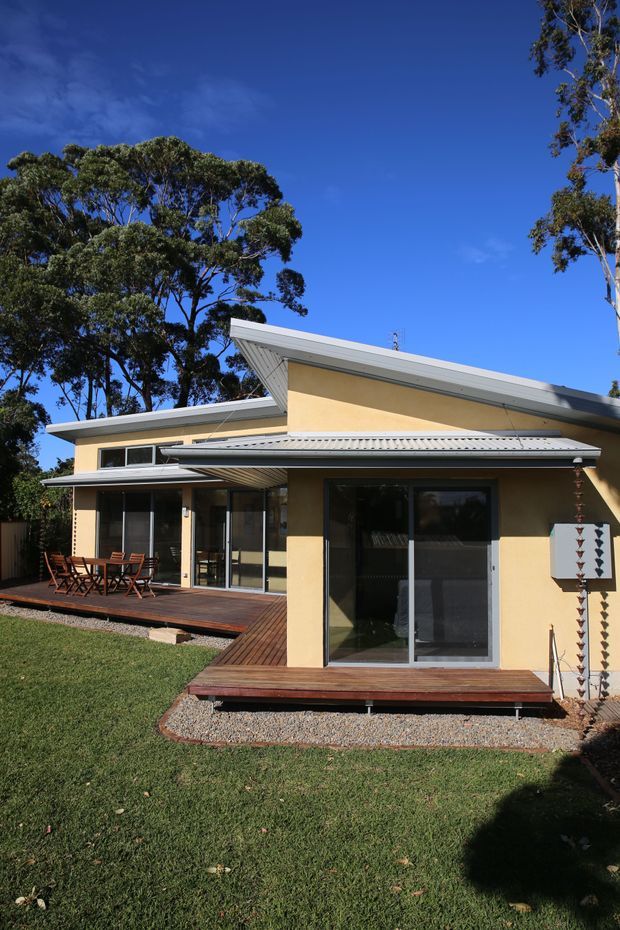Exterior view of a single-storey suburban home with pale rendered walls, multiple low-pitched skillion roof planes, broad eaves, large glazed sliding doors, and timber decking extending into a grassed yard. Mature trees surround the house under a clear blue sky.