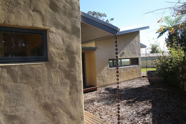Exterior view of a low-profile modern residence with textured rendered walls, dark-framed horizontal windows, a projecting metal roof and sheltered entry. A timber deck and landscaped gravel courtyard with a decorative rain chain are visible in the foreground.