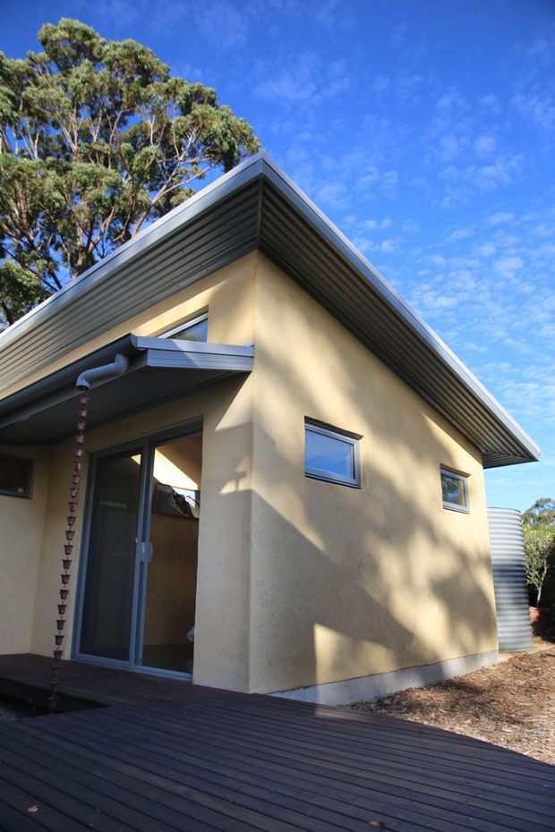 Exterior view of a compact single-storey house with a steeply pitched gable roof, dark metal fascia and soffit, pale rendered walls, narrow horizontal windows, and a glazed door opening onto a dark timber deck. A corrugated cylindrical water tank is visible beside the building, with mature trees surrounding the site.