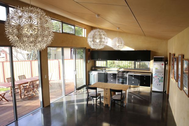 Open-plan kitchen and dining area with high ceilings, extensive glazing and sliding doors opening to a timber deck. The room features polished dark concrete flooring, warm timber wall and ceiling finishes, black cabinetry, stainless-steel appliances, a timber dining table, and sculptural spherical pendant lights.