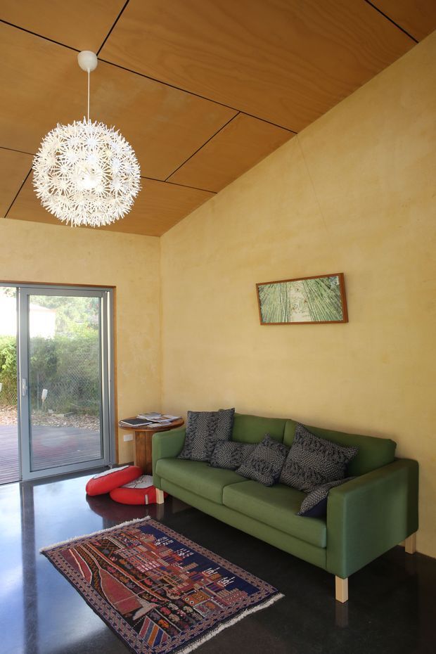 A compact, warmly styled living room with ochre plaster walls, a richly grained timber-panelled ceiling, and a large white spherical pendant light. A green sofa with patterned cushions sits beside a small timber side table, with a colourful patterned rug over a dark polished floor. Full-height glazed doors open to a timber deck and garden, while a framed botanical artwork hangs above the sofa.