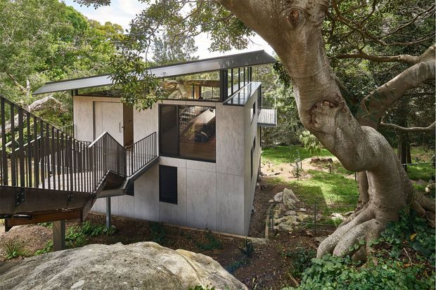 Exterior view of a compact contemporary multi-level house or studio set among mature trees and natural rock. The building has a sharply sloping roof, pale panelled walls, extensive dark-framed glazing, and black metal stairs and balustrades connecting elevated levels. The structure appears carefully integrated into a leafy, sloped landscape.