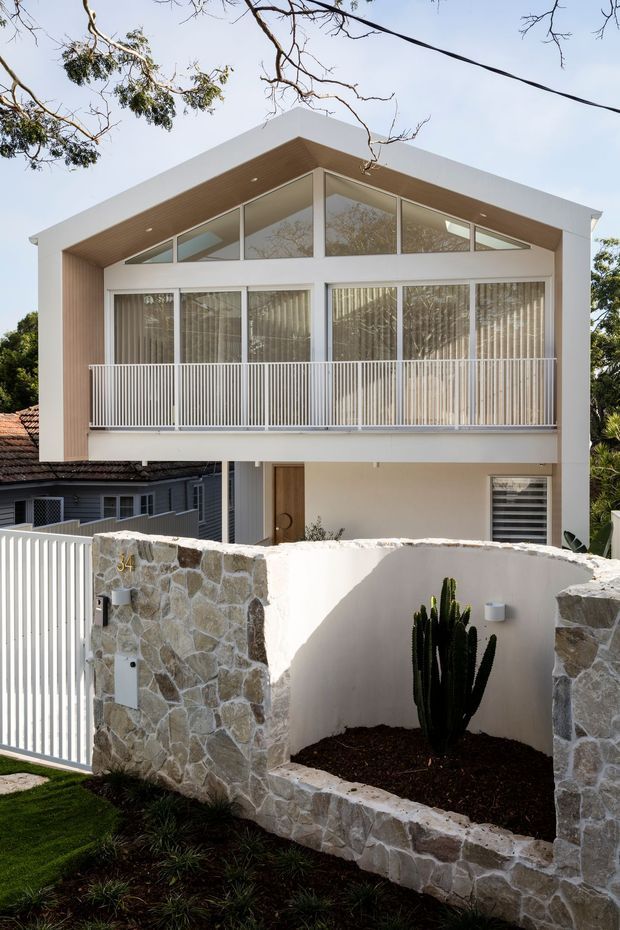 Front exterior view of a contemporary multi-level residence with a prominent pitched gable roof, white rendered facade, extensive upper-level glazing, a covered balcony with slim vertical metal balustrades, and warm timber soffit lining. The property is enclosed by a rough natural-stone and rendered masonry wall with an integrated cactus garden and white pedestrian gate.