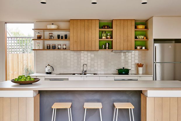 Bright, modern kitchen featuring a broad island with three stools, pale timber vertical-panel cabinetry, open shelving, a light tiled splashback, white benchtops, integrated appliances, and a stainless-steel refrigerator. A glazed opening brings in natural light and provides a view to an exterior fence and greenery.