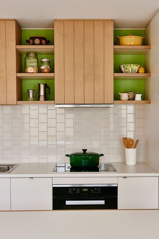 Compact, brightly styled kitchen with light timber cabinetry, vivid green open-shelf backing, white glossy brick-pattern tiled splashback, pale benchtops, and integrated black oven and induction cooktop. Open shelves display jars, cookware and ceramics, while a green enamel pot provides a strong accent.