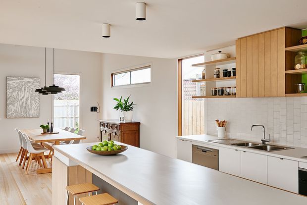 Bright open-plan kitchen and dining area with a large pale island, timber stools, white cabinetry, vertical timber wall cabinetry, and a white rectangular tile splashback. The kitchen opens toward a dining space with a timber table and white chairs, while natural light enters through multiple windows and glazing.