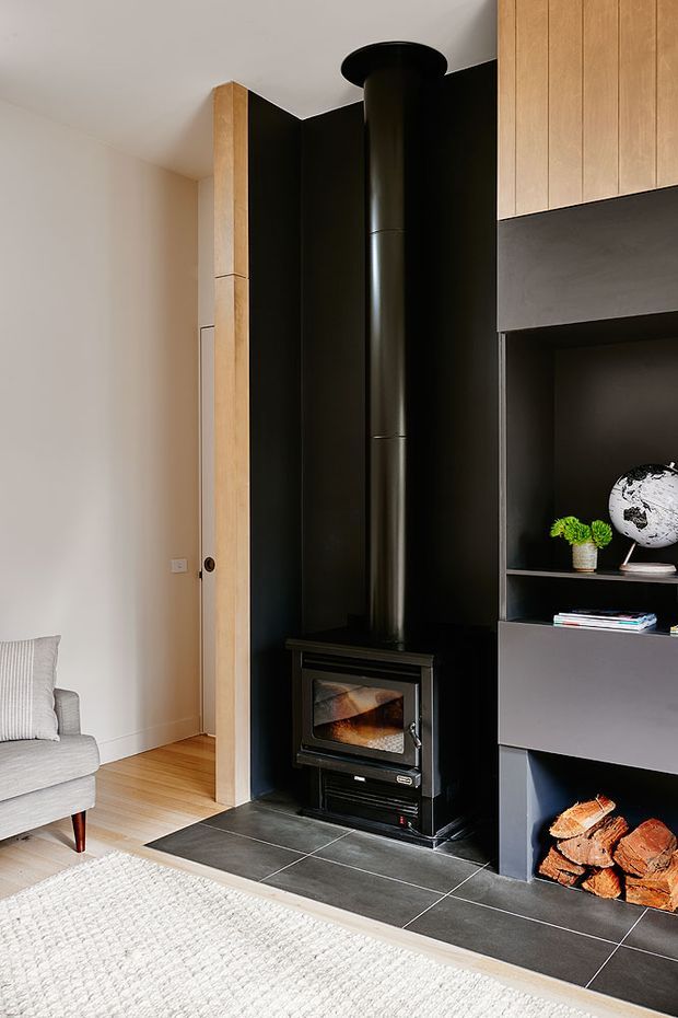 A contemporary living room featuring a freestanding black wood-burning stove with a tall flue set against a dramatic black tiled feature wall. Built-in dark shelving and cabinetry frame the fireplace area, with warm timber cabinetry and trim providing contrast. A light upholstered armchair, textured rug, firewood storage, and small decorative objects complete the refined interior.