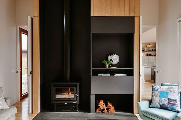 Interior view centered on a dark built-in fireplace and media/storage wall. The composition combines black and charcoal cabinetry with warm vertical timber panelling and a tiled hearth floor. A freestanding wood-burning stove sits to the left, while an open firewood niche and display shelving occupy the lower and central sections. A blue upholstered armchair is visible at right, with a hallway or adjacent room beyond.