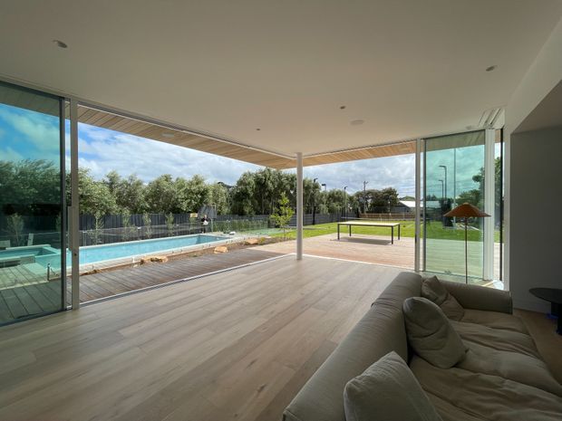 A bright, open-plan living room with a large neutral sectional sofa and extensive floor-to-ceiling sliding glass doors opening onto a timber deck and landscaped backyard. A swimming pool, lawn, outdoor table-tennis table, and covered outdoor area are visible beyond the glazing. The interior has pale timber flooring, a white ceiling with recessed downlights, and a contemporary indoor-outdoor connection.