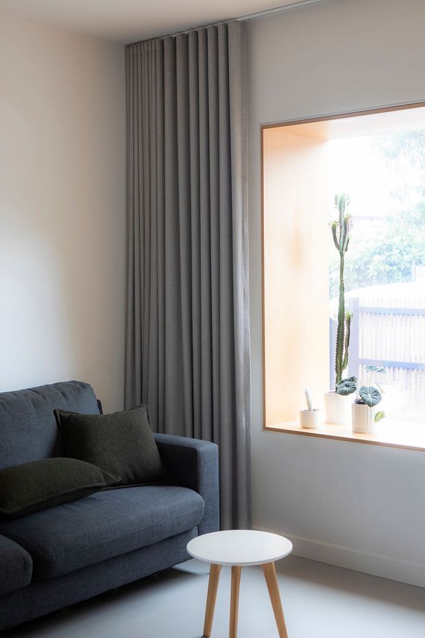A bright, minimalist living room corner with a dark charcoal upholstered sofa and cushions, a small round white coffee table with timber legs, full-height dark grey curtains, and a large timber-framed window with potted plants on the sill. The room has pale walls, a light smooth floor, and abundant natural light.