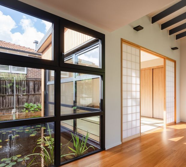 Bright contemporary interior corridor or living space with warm timber flooring, black-framed floor-to-ceiling glazing, and a view into a planted courtyard with a small pond. A timber-trimmed shoji-style sliding partition opens to a light-filled adjoining room, while exposed dark ceiling beams add contrast.