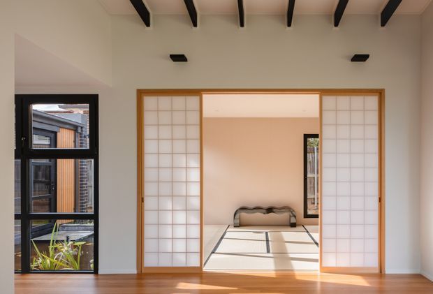 A bright contemporary interior looking through timber-framed shoji-style sliding screens into a tatami-floored room. The foreground has warm timber flooring, white walls, exposed dark ceiling beams, and a black-framed glazed door or window at left. The adjoining room contains a low bench, pale tatami mats, and a small dark-framed window, creating a restrained Japanese-inspired atmosphere.