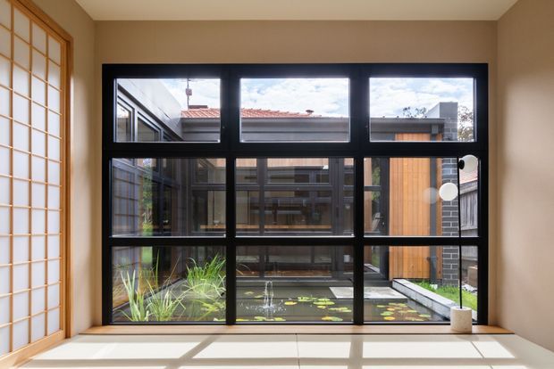 Interior view through a large black-framed multi-pane window overlooking a landscaped courtyard with a shallow pond and contemporary timber-and-brick building elements. A timber-framed translucent sliding door or screen is visible at left, while warm beige walls and pale tiled flooring create a calm, sunlit setting.