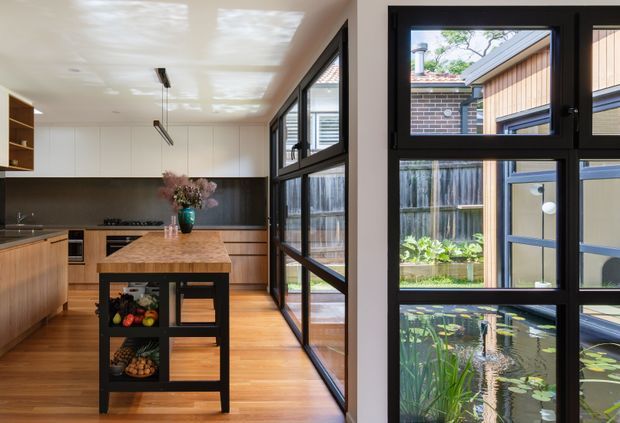 A contemporary kitchen viewed toward a glazed courtyard connection. The room features warm timber flooring and cabinetry, a stone island with integrated dark shelving, black-framed full-height windows and doors, and a minimalist white ceiling. The glazing overlooks a landscaped courtyard with a water feature and an adjacent timber-clad structure.