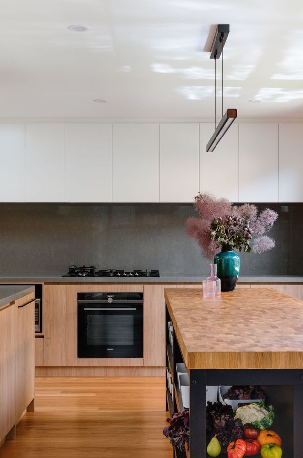 A contemporary kitchen featuring full-height white overhead cabinetry, a dark stone or concrete-look splashback, timber cabinetry and flooring, and a large timber butcher-block island with dark framing. A gas cooktop and built-in oven line the rear wall, while a linear pendant light hangs above the island. Decorative dried flowers and produce add colour to the otherwise warm neutral scheme.