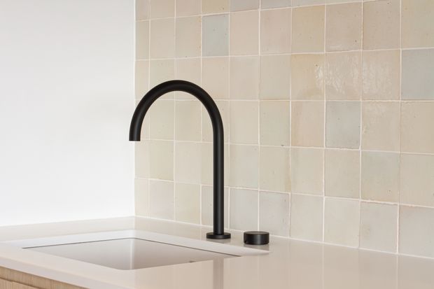 Close-up of a minimalist kitchen sink area with a pale undermount sink, tall matte black curved tap and separate black control, set against a handmade-looking square tiled splashback in soft cream, beige and muted grey tones.