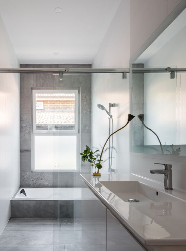 A bright, compact contemporary bathroom with a bathtub beneath a frosted lower window, a clear glass shower screen, and a long white integrated vanity with chrome tapware. Large grey stone-look tiles cover the floor and rear wall, while a full-width mirror reflects the shower and plant. The room has clean lines, recessed ceiling lights, and a restrained white, grey, and chrome palette.