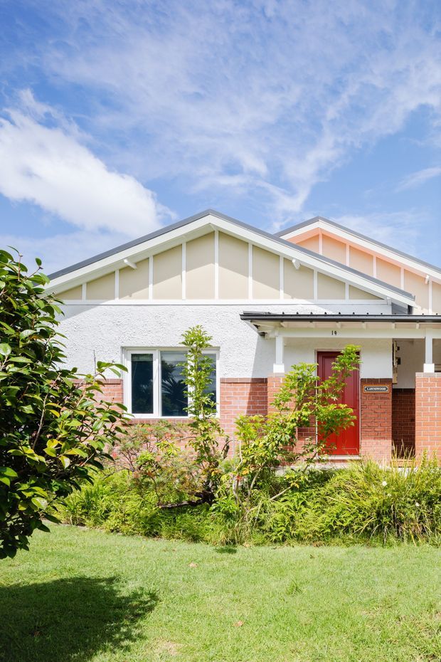 A bright exterior view of a single-storey suburban home with a prominent gabled roof, white textured rendered walls, exposed decorative gable framing, red brick lower walls, and a red front door beneath a covered entry porch. The house is set behind a landscaped front garden and lawn under a blue sky.