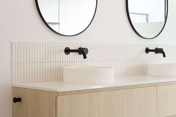 Contemporary bathroom vanity with a pale timber floating cabinet, white integrated benchtop, round vessel basins, matte black wall-mounted tapware, and a vertically ribbed light tile splashback. Two large round black-framed mirrors sit above the double vanity, creating a clean, softly minimal look.