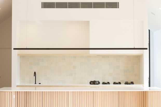 Bright contemporary kitchen with a long island featuring vertically fluted timber cabinetry, a pale stone benchtop, a softly variegated light tile splashback, and a linear black pendant light. The rear wall includes a concealed rangehood vent, sink, and four-burner gas cooktop, with minimal white cabinetry and clean architectural lines.