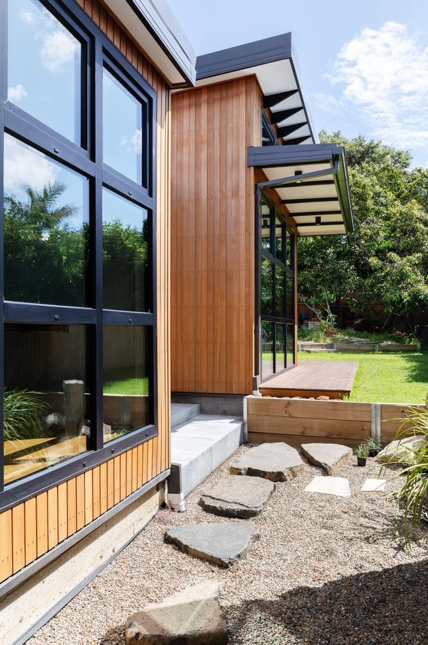 Exterior view of a contemporary timber-clad house with extensive black-framed glazing, projecting flat-roofed covered deck, timber decking, concrete steps, and a gravel garden path with irregular stone pavers. The building opens onto a landscaped backyard with lawn and established planting.