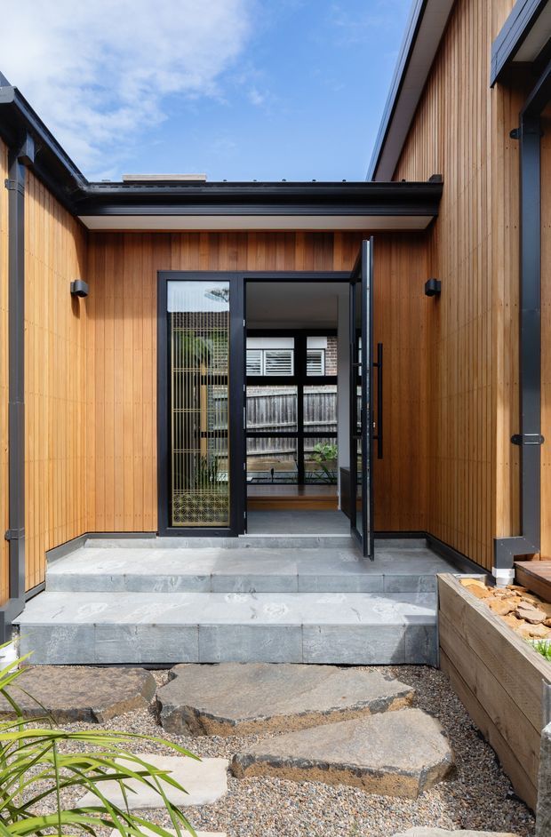 Exterior view of a contemporary residential entry courtyard. Warm vertical timber cladding surrounds a black-framed glazed doorway, with broad grey stone steps leading up from an informal gravel and stone path. The partially open door reveals a glazed interior and planted courtyard beyond. Dark metal roof edges, gutters and trims create strong contrast against the timber facade.