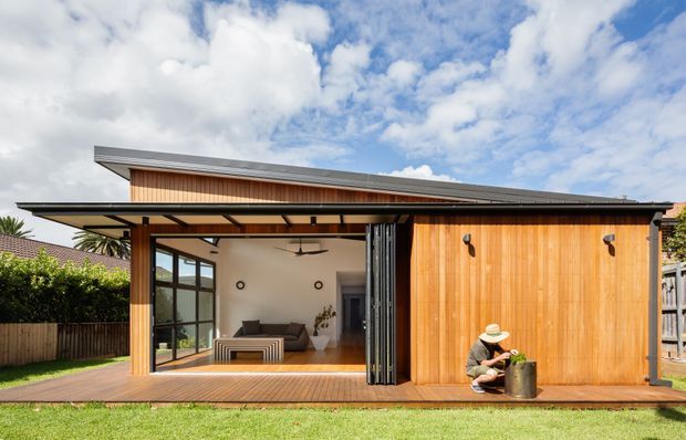 Exterior view of a contemporary single-storey timber-clad pavilion or extension with a low-pitched asymmetrical roof, extensive opening glazed sliding doors, and a covered timber deck. The open living area is visible inside, while a large timber-clad wall encloses the right side. The building sits in a landscaped suburban garden under a bright, partly cloudy sky.