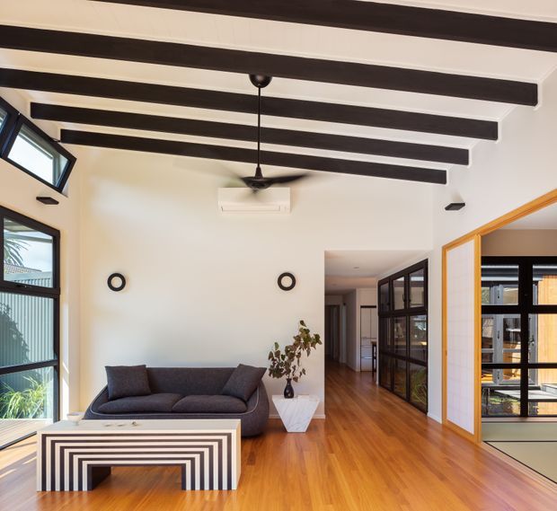 Bright contemporary living room with warm timber flooring, white walls, exposed dark ceiling beams, large black-framed windows, and a central ceiling fan. A charcoal sofa and bold geometric coffee table occupy the foreground, while a glazed internal partition and timber-framed opening connect to adjacent spaces.
