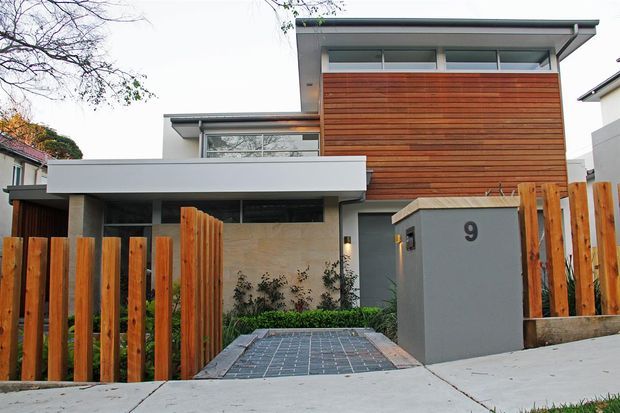 Street-facing exterior of a contemporary multi-level residence with a flat roof, projecting white roof forms, extensive horizontal timber cladding, grey rendered walls, and a paved entry driveway. Vertical timber fencing and a numbered grey entry pillar frame the frontage.