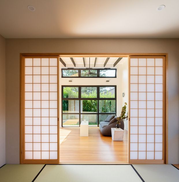 View from a tatami-floored room through timber-framed shoji sliding doors into a bright contemporary living area. The adjoining room has warm timber flooring, extensive dark-framed glazing, exposed ceiling beams, a grey sofa, and potted greenery, with a garden visible beyond.
