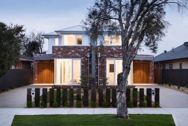 Front exterior view of a contemporary two-storey residence with a symmetrical composition, exposed red brick upper and central facade, pale metal roof, large glazed openings, and warm timber garage or entry doors. A mature street tree, low timber-and-planting fence, paved driveways, and manicured lawn frame the house.