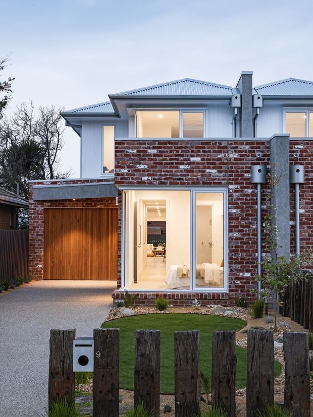 Exterior view of a contemporary multi-level townhouse or residential extension, featuring a red exposed-brick lower facade, pale metal-clad upper level with pitched roof, timber garage door, large glazed openings, and a compact landscaped front yard with timber posts.