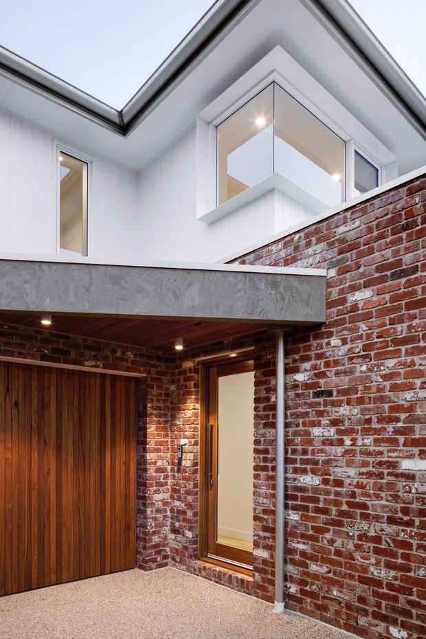 Exterior architectural view of a compact courtyard or covered entry, combining retained red brick walls with a contemporary white upper-level extension. The upper facade features white vertical cladding, angular roof forms, and projecting corner glazing. Below, a timber garage door, timber-framed entry door, exposed brickwork, concrete beam, and timber-lined soffit create a warm material contrast. The space appears bright and carefully detailed, with a narrow courtyard-like forecourt.