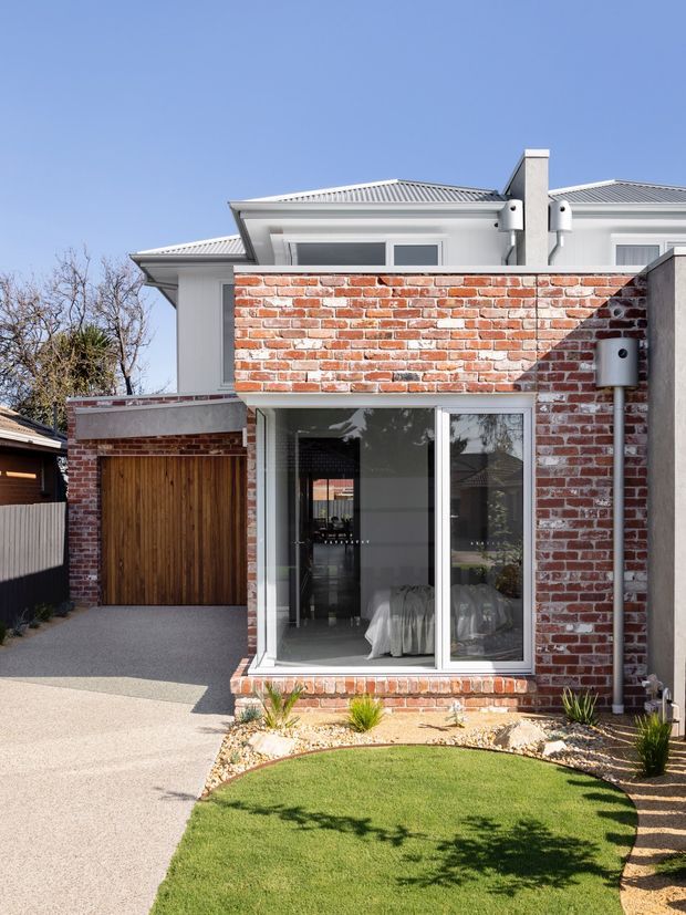 A bright exterior view of a contemporary multi-level residential extension. The facade combines exposed red brickwork, smooth light-coloured walls, large white-framed glazing, and a corrugated metal hipped roof. A timber garage door, paved driveway, small landscaped lawn, and gravel garden border are visible.