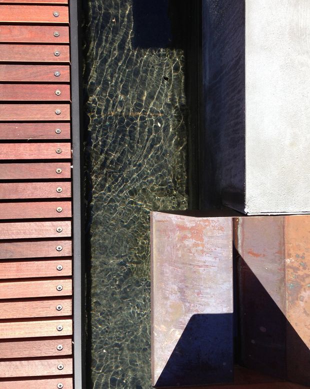 Close-up architectural detail showing a narrow channel of dark rippling water or reflective glazed surface between reddish timber slatted screening on the left and a dark wall with weathered terracotta or rust-toned cladding on the right. Strong sunlight creates graphic shadows and highlights the contrasting linear, textured materials. No room interior is visible.