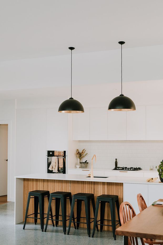 Bright contemporary kitchen featuring a long white island with a vertical timber slat front, four dark bar stools, white flat-panel cabinetry, a fine white tiled splashback, integrated oven, black cooktop and a pale stone or terrazzo-style floor. Two large dark pendant lights hang above the island, while a dining table and chairs are partially visible in the foreground.