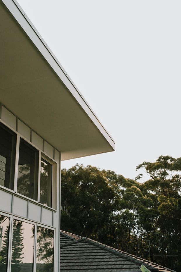 Close-up exterior architectural view of a modern, box-like building with a broad projecting flat roof, pale metal-framed floor-to-ceiling glazing, and light grey panelled cladding. A tiled pitched roof of an adjacent structure and mature trees are visible in the background.