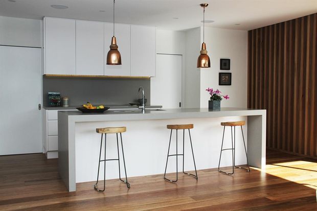 A bright, minimalist contemporary kitchen with white flat-panel cabinetry, a long white island with integrated seating, dark timber flooring, copper pendant lights, and a full-height vertical timber feature wall. Three bar stools line the island, while daylight enters from the right.