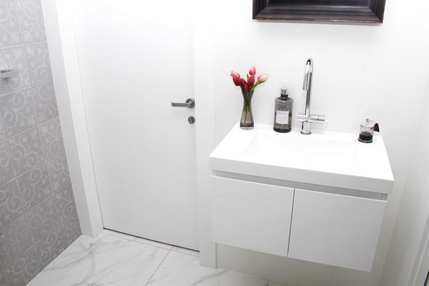 Compact contemporary bathroom vanity area with a floating white cabinet, white benchtop, chrome wall-mounted tapware, frameless mirror edge, and white marble-look floor tiles. A patterned grey feature wall and a closed white door are visible, with small decorative accessories on the vanity.