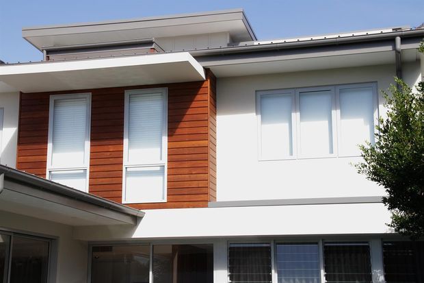 Exterior view of a contemporary multi-level residential building with white rendered walls, warm horizontal timber cladding, broad flat roof overhangs, and a mix of tall narrow and wide windows. The facade is photographed in bright daylight and includes a lower-level glazed area.