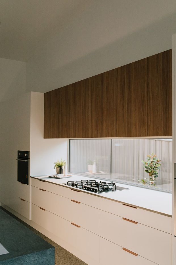 A contemporary galley-style kitchen with extensive pale cabinetry and integrated timber-grain overhead cupboards. The white benchtop includes a four-burner gas cooktop, with a large glazed window and sheer curtains behind it. A dark stone or tiled island edge is visible in the foreground, complemented by warm wood cabinetry and minimal linear handles.