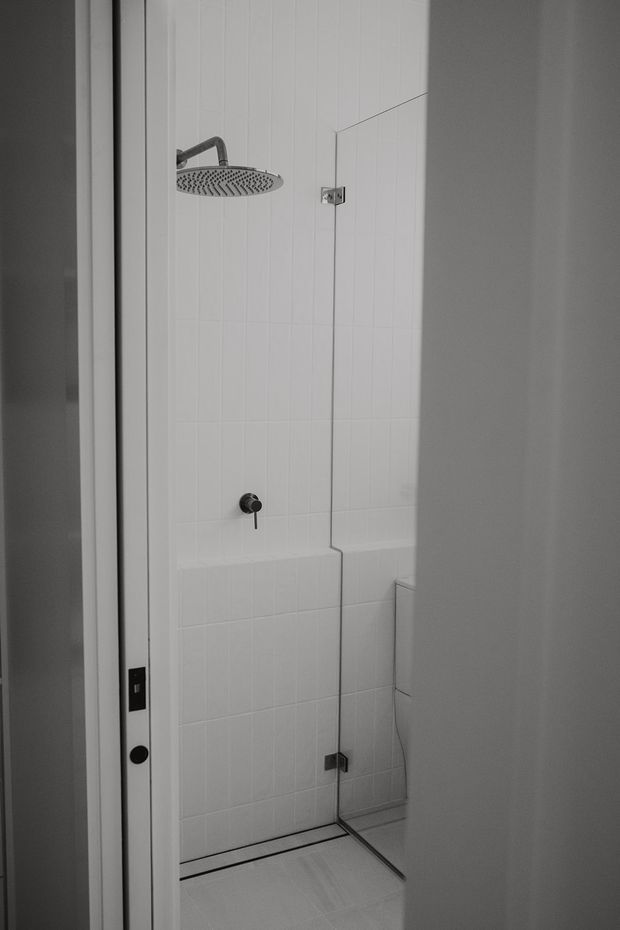 A narrow contemporary walk-in shower enclosure viewed through a partially open door. The shower has white tiled walls, a tiled floor, a large round rainfall showerhead, dark shower controls, and clear frameless glass panels with minimal hardware.