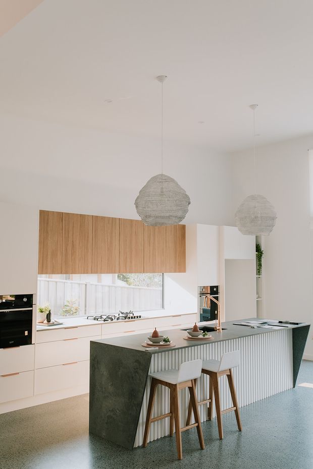 Bright contemporary kitchen with a large island, white flat-panel cabinetry, warm timber overhead cupboards, a dark stone waterfall benchtop, and a vertically fluted island front. Two woven pendant lights hang over the island, which is styled with stools and place settings. Large windows provide natural light, while integrated appliances and open shelving complete the clean, refined fit-out.