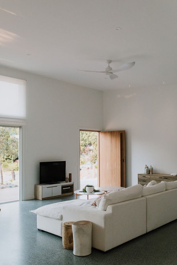 Bright, minimalist living room with a large cream sectional sofa facing a television, white walls and ceiling, a pale terrazzo-like floor, timber pivot door, and generous glazing opening to greenery. A white ceiling fan and simple low timber media console complete the calm, contemporary interior.