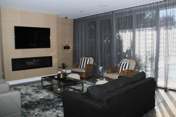 Contemporary living room with a dark sectional sofa, two wicker lounge chairs, a glass coffee table, patterned rug, wall-mounted television and built-in linear fireplace. A full-height glazed wall with sheer curtains brings in filtered daylight, while the pale stone or wood-look feature wall creates a refined focal point.